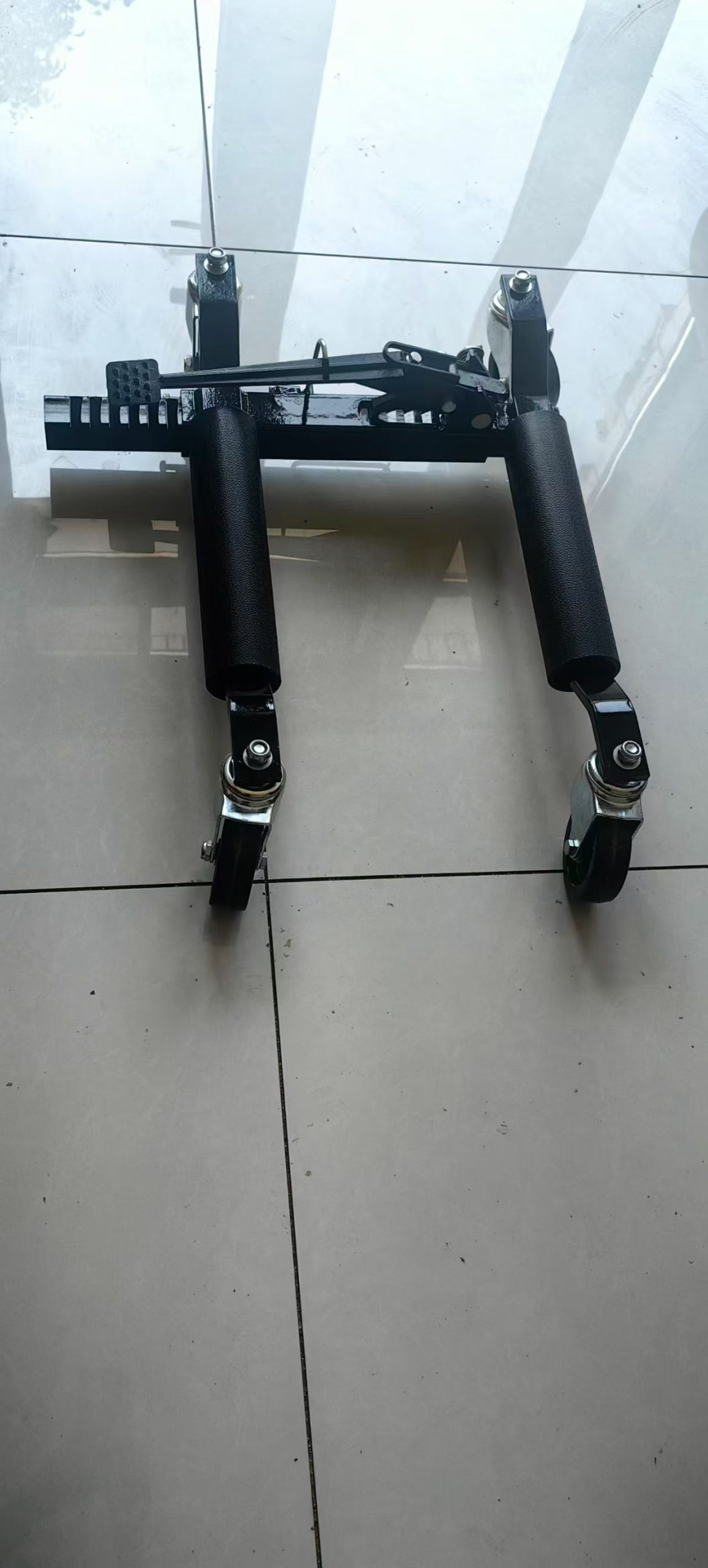 Top view of a black wheel dolly showing pedal, toothed bar and twin rollers
